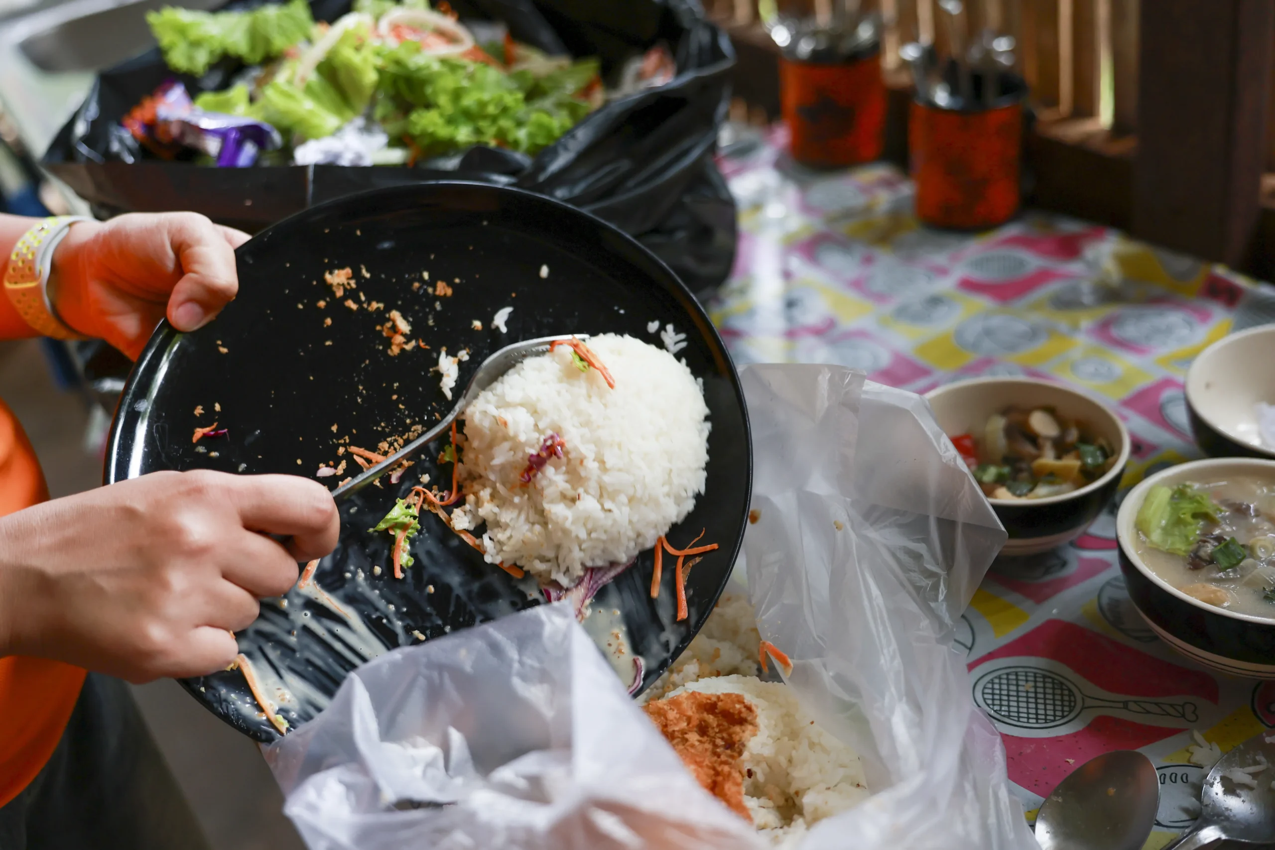 A person scraping leftover rice and food from a black plate into a clear plastic garbage bag.