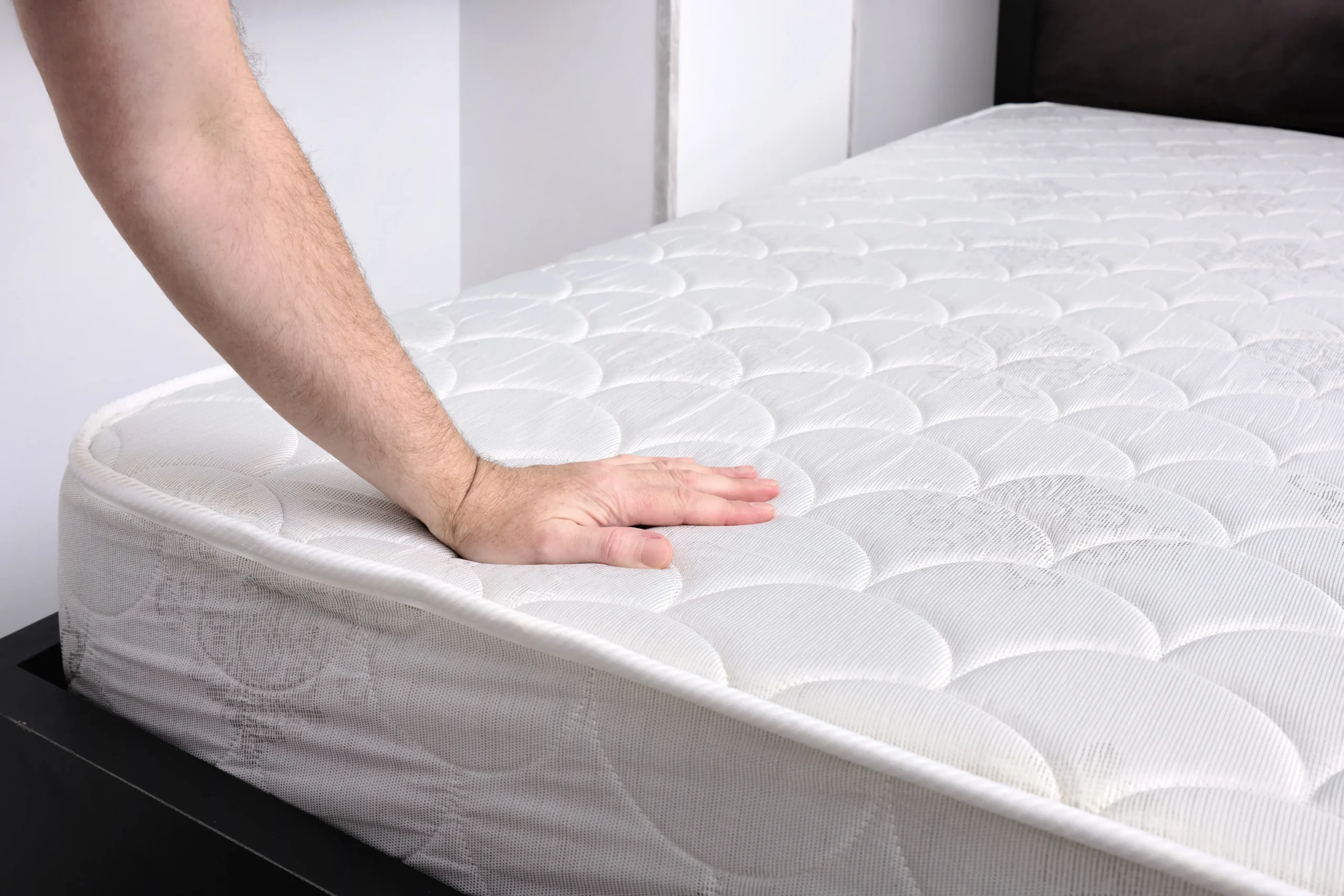 A person's hand pressing down on a bare, white quilted mattress while inspecting it for a bed bug infestation.