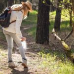 Woman on a wooded trail applying bug spray to pant leg.